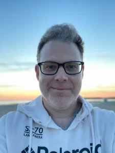 Color photograph of Bill Wolff, a white man with dark rimmed glasses, at sunset, wearing a white hoodie with the hood down. He is looking directly at the camera with a kind of smirk on his face. The sky goes from light blue to white to yell to orange in the background.